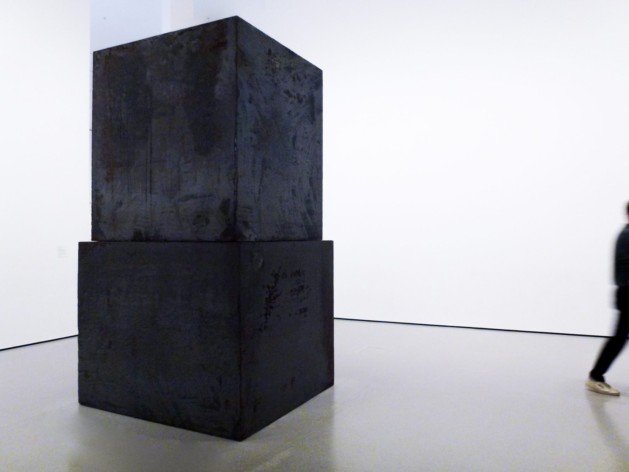 A dark stacked-cube sculpture in a white gallery as a visitor walks past